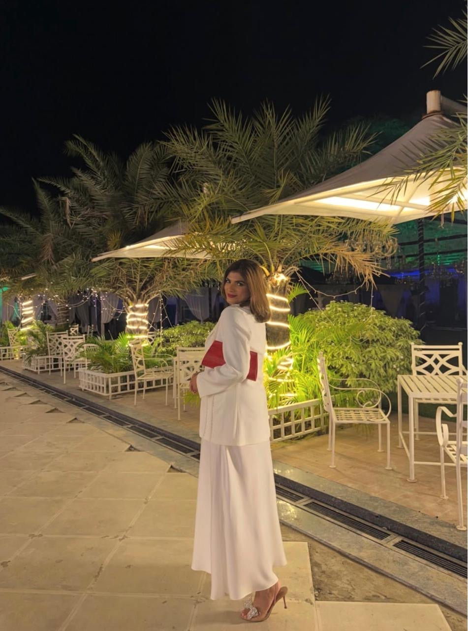 A woman wearing a white traditional outfit with red accents and nude heels stands on a patio at night, with illuminated palm trees and white outdoor furniture in the background.