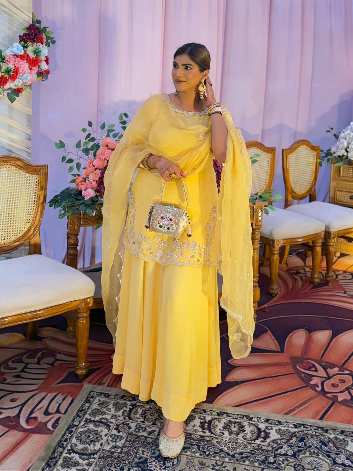 A woman wearing a bright yellow traditional outfit with a matching sheer dupatta and a small patterned clutch bag, standing in an indoor decorated event space with pink drapery, floral arrangements, chairs, and a patterned rug.